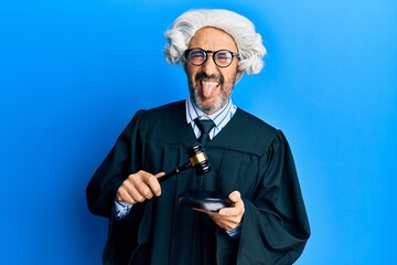 Middle age hispanic man using gavel sticking tongue out happy with funny expression.