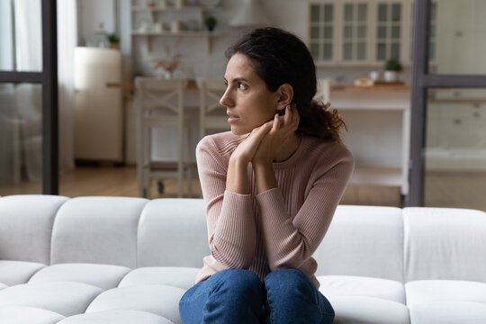 Thoughtful young hispanic woman sitting on sofa in living room, looking in distance, considering problem solution alone indoors. Nervous millennial lady thinking on personal psychological troubles.