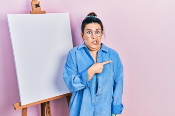 Young hispanic woman standing by painter easel stand pointing aside worried and nervous with...