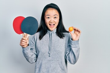 Young chinese girl holding red ping pong rackets and ball celebrating crazy and amazed for success...