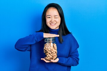 Young chinese girl holding jar of chocolate chips cookies winking looking at the camera with sexy...