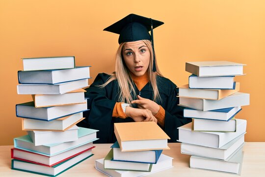 Young caucasian woman wearing graduation ceremony robe sitting on the table in hurry pointing to watch time, impatience, upset and angry for deadline delay