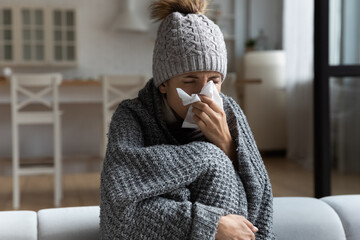 Unhealthy young woman in warm clothes using paper tissue, suffering from runny nose, caught cold,...