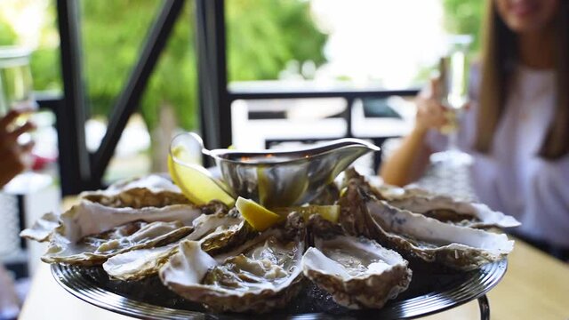 Luxury restaurant visitors clinking glasses with wine and cheering behind served plate of oysters on ice with lemon, sauce. Salver tray of traditional meditarian dish sea food for gourmets on a table.