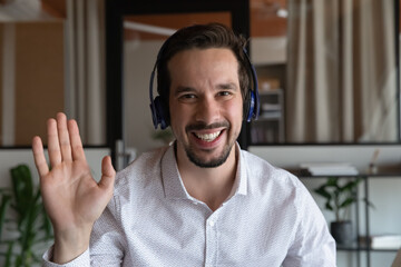 Head shot portrait of smiling man in wireless headphones waving hand and looking at camera, business mentor coach teacher greeting students viewers, recording webinar, employee making video call