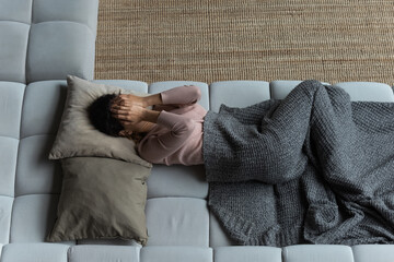 Above top view unhappy frustrated young woman lying on sofa on pillow under plaid, covering face...