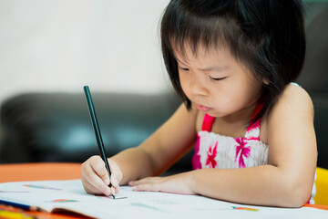 A 3-4 year old Asian child girl is doing her homework. Kid hold black crayons to paint in their workbooks. Children learn at home.