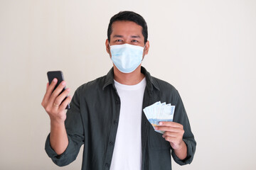Adult Asian man wearing protective mask while holding mobile phone and paper money