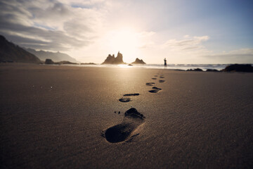 Footprints in sand against silhouette of person. Man walking along beach to sea at golden sunset....
