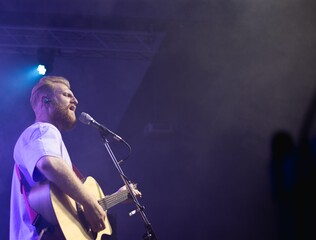 A young man with a beard holds an acoustic guitar in his hand and sings into a microphone stands on...