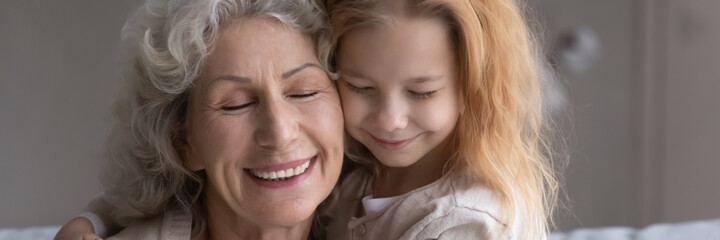 Happy faces of excited older grandmother and little preteen girl granddaughter cuddling after long separation touch cheeks with closed eyes feel love affection. Panoramic header image website banner