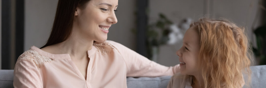 Talking mother and child. Happy single foster mom sit on couch speak to small junior school age girl adopted daughter enjoy conversation discuss mutual interests. Panoramic banner image for website
