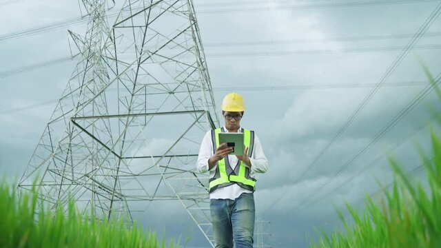 Font View.Electrical engineer wearing a Yellow helmet and safety carrying using tablet vest walking near high voltage electrical lines towards power station on the field..