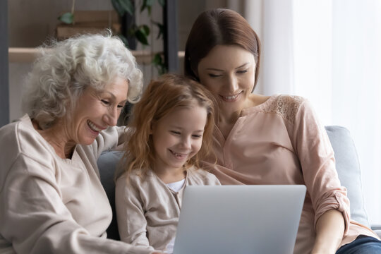 Three age family. Happy little girl daughter grandkid sit on sofa between elderly grandma and adult mom watch video movie on laptop. Smiling females of diverse generations surf internet on pc together