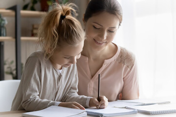 Out of school education. Helping young mom engaged in learning activity with preteen daughter assist in studying maths or english rules. Smiling millennial lady teacher give home lesson to little girl