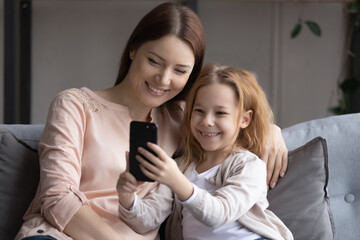 Smiling junior school age girl sit on sofa with young adult mother use smartphone take selfie for...