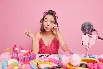 Fototapeta premium Woman video channel host skilled cosmetologist uses eyelashes curler tells about applying makeup presents cosmetics onine wears hair rollers pink dress poses indoor. Blogging and freelancing concept