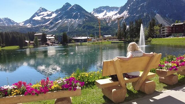 Woman tourist relaxing on a park bench of Arosa town and tourist resort by Obersee Lake in Switzerland. Arosa lakefront by cable car station to Aroser Weisshorn peak. Plessur Region in Grisons Canton.