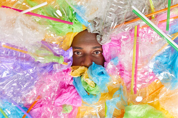 Serious black man covered with plastic garbage shows half of face considers pollution problem...