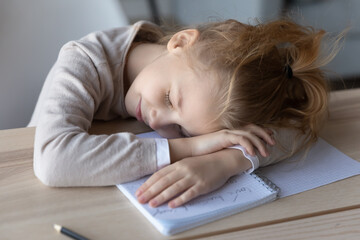 Boring lesson. Tired little girl lying on work desk put head on opened copybook sleep nap lose...