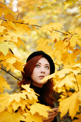 Naklejka na ściany i meble Cute girl in yellow foliage looks up. The fall period trees. Autumn romantic portrait of a red-haired woman. Walk in the Park.
