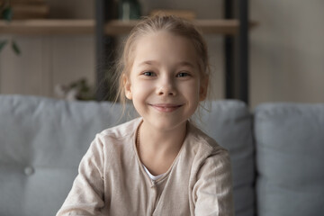 Headshot webcam portrait of smiling little girl junior school age sit on couch at home look at...