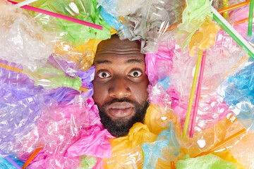 Black man sticks head in plastic garbage has stunned expression thick beard bothered with problem of trash surounded by crumpled bottles straws and colorful polythene bags. Danger for ecology