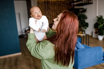 Joyful young Caucasian red haired mother lifting cute baby son, playing with biracial african...