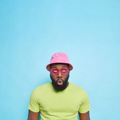 Impressed surprised dark skinned guy with thick beard focused above keeps mouth opened wears panama casaul green t shirt heart shaped sunglasses isolated over blue background with blank space