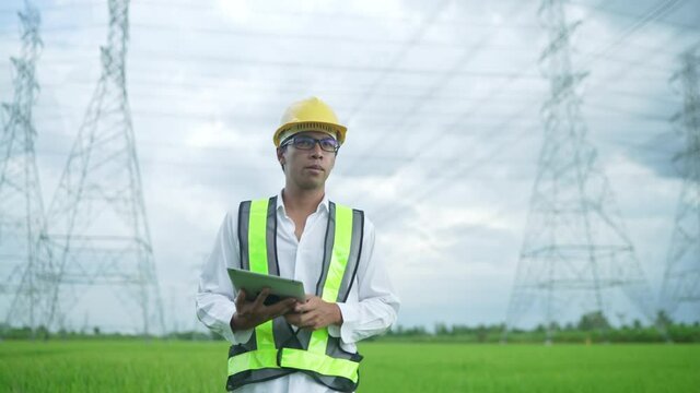 Font View.Electrical engineer wearing a Yellow helmet and safety carrying using tablet vest walking near high voltage electrical lines towards power station on the field..
