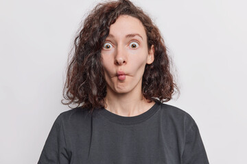 Pretty surprised young female model makes grimace folds lips looks with shocked expression at camera has dark wavy hair wears black t shirt isolated over white background does facial exercises