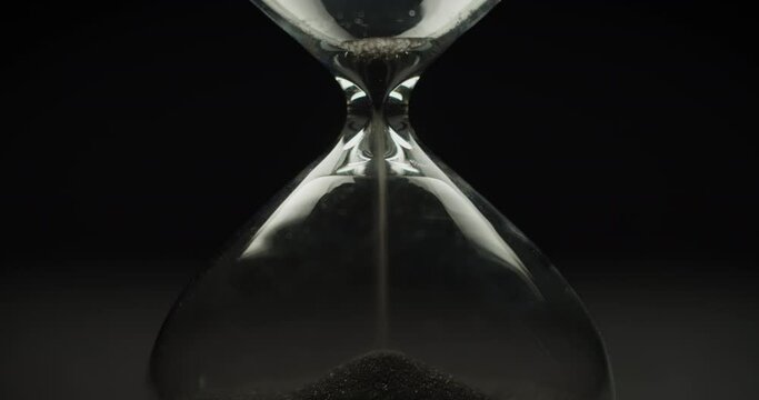 Black sand pouring inside the modern hourglass on a black background. The end of time cycle. Close up. 
