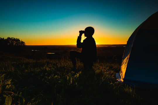 Silhouette of a man with binoculars near the tent against the backdrop of the sunset on a clear sky over the meadow.