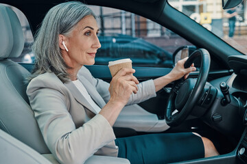 Mature beautiful woman in smart casual wear smiling and drinking coffee while driving car