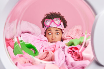 Stunned worried young African American housewife has scared expression hides in pile of laundry poses from inside of washer wears snorkeling glasses on forehead busy with everyday domestic work