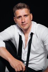 A stylish young man in a white shirt is sitting on the director's chair on a black background. A handsome charismatic businessman.