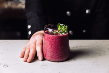 Bartender holding a glass of refreshing forest fruit berry smoothie cocktail garnished with a mint sprig, marble table.