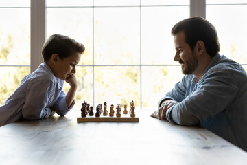 Focused thoughtful clever son and dad playing chess, sitting at table with chessboard, thinking...