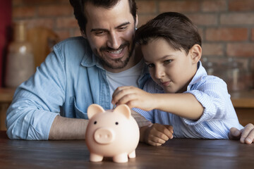 Happy dad teaching smart little son to save and invest money, encourage kid for accounting, investment, planning future budget. Boy hugging father, dropping cash into piggy bank. Family economy - obrazy, fototapety, plakaty