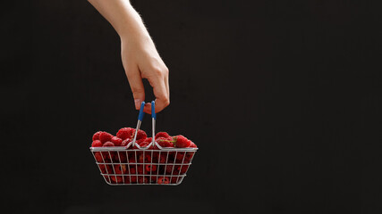 Girl's hand holding a mini grocery shopping basket with fresh raspberries on a dark background