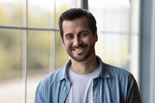 Portrait of happy handsome millennial young 30s business man, teacher, entrepreneur wearing casual, standing at window in apartment, looking at camera, smiling. Head shot, video call screen view