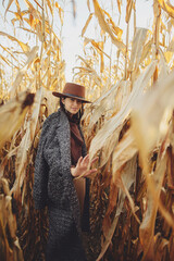 Happy stylish woman in brown hat and vintage coat walking in autumn maize field in sunset light. Fashionable young hipster female in retro outfit smiling in autumnal corn