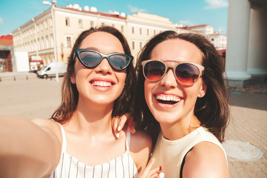 Two young beautiful smiling hipster female in trendy summer clothes.Sexy carefree women posing on the street background in hat. Positive pure models having fun at sunset, taking Pov photos in the city