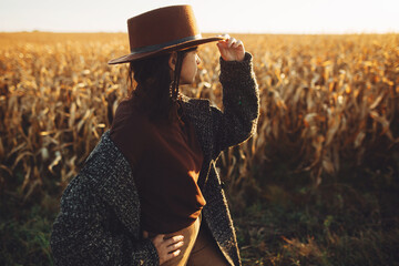 Beautiful stylish woman in brown hat walking in sunset light in autumn field. Atmospheric moment. Fashionable young hipster female in retro outfit standing at maize field in evening countryside