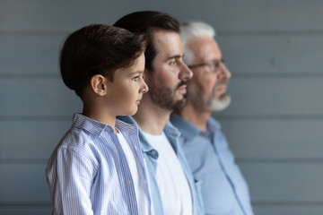 Serious thoughtful cute boy kid standing in row with young father and senior grandfather, looking...