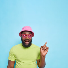 Handsome happy African American man with thick beard points at upper right corner dressed in summer clothes trendy pink sunglasses shows copy space for your advertisement isolated on blue background
