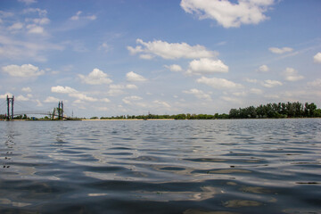 Spring scenery of a reflective surface of a lake, blue cloudy sky and distant bridge with green line of trees