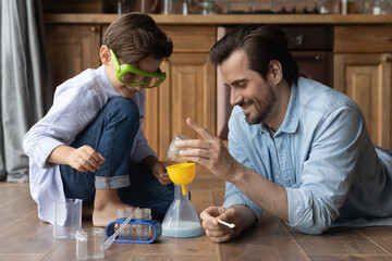 Happy dad and cute preschooler kid engaged in learning chemical game, resting on heating floor at home, imitating experiment in chemistry, studying science, playing with beakers, pouring liquids - obrazy, fototapety, plakaty