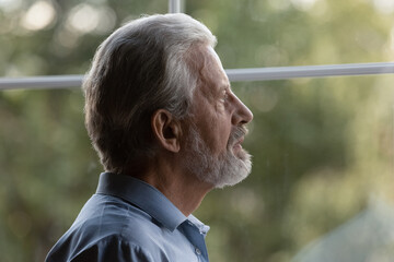 Sad pensive older senior 70s man lost in thoughts looking out window, suffering from memory loss,...