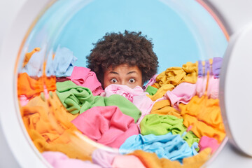 Shocked troubled ethnic woman hides behind big pile of laundry oveloaded with housework and domestic responsibilities stares bugged eyes poses from washing machine drum. Deadline for washing